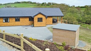A wooden building with a grey roof, a covered outdoor seating area, and a fenced garden. - Woodland Lodge - UK6651 (Hundred House, near Builth Wells)