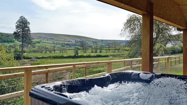 A hot tub with a wooden deck and railing, overlooking a green landscape with hills and trees. - Woodland Lodge - UK6651 (Hundred House, near Builth Wells)
