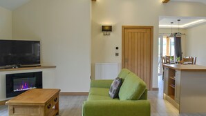 A living room with a flat-screen TV, a fireplace, a wooden coffee table, a green sofa, and a dining area with a wooden table and chairs. - Woodland Lodge - UK6651 (Hundred House, near Builth Wells)