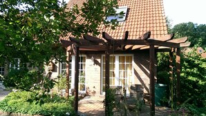 Une maison en briques avec une pergola en bois, une terrasse en briques et un jardin où poussent différentes espèces de plantes.