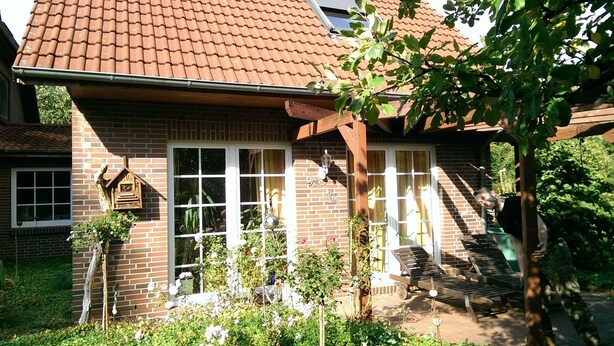 Une maison en briques avec un toit en tuiles, un porche à l’avant avec deux chaises en bois et un petit jardin agrémenté de diverses plantes et fleurs.