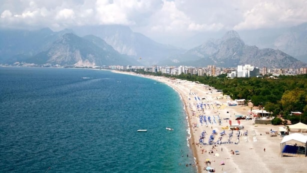 A coastal cityscape with a beach, mountains in the background, and a clear sky. - Karden Otel (Antalya)