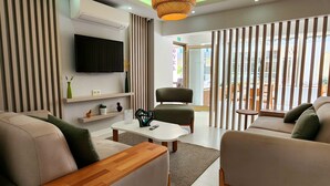 A modern hotel lobby with a sofa, a wooden coffee table, a television, and a large window with vertical blinds. - Karden Otel (Antalya)