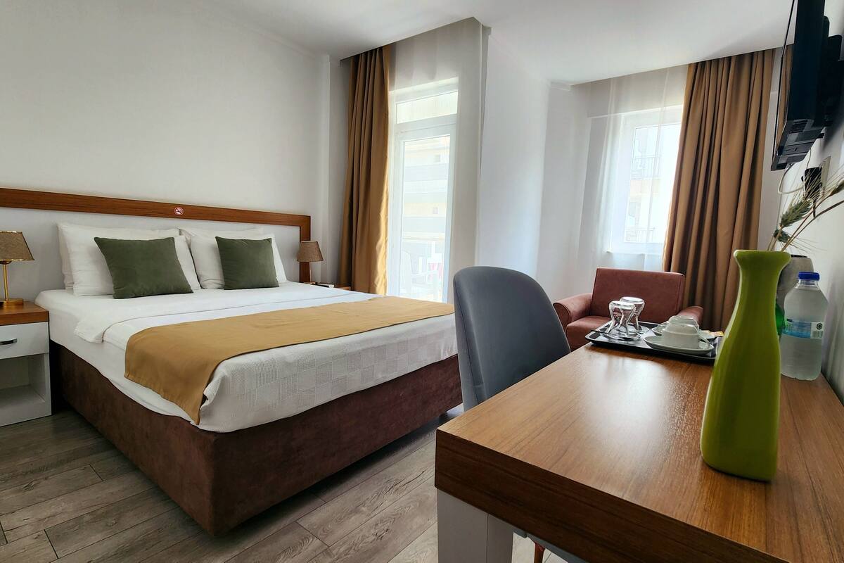 A hotel room with a bed, bedside table with a lamp, a desk with a chair, a water bottle, and a vase.