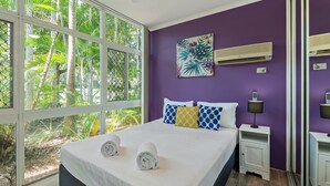 Une chambre à coucher comprenant un lit, deux tables de chevet, un climatiseur fixé au mur et une fenêtre donnant sur un paysage verdoyant.