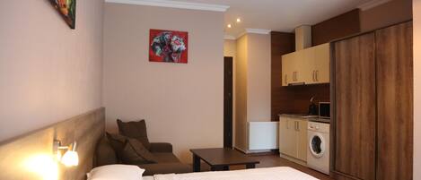 A bedroom with a bed, a nightstand, a chair, a desk, a painting on the wall, and a kitchen area with a washing machine.