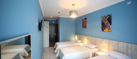 Quarto de hotel com quatro camas de solteiro, cabeceira de madeira, televisão fixada na parede e porta de acesso a um banheiro.