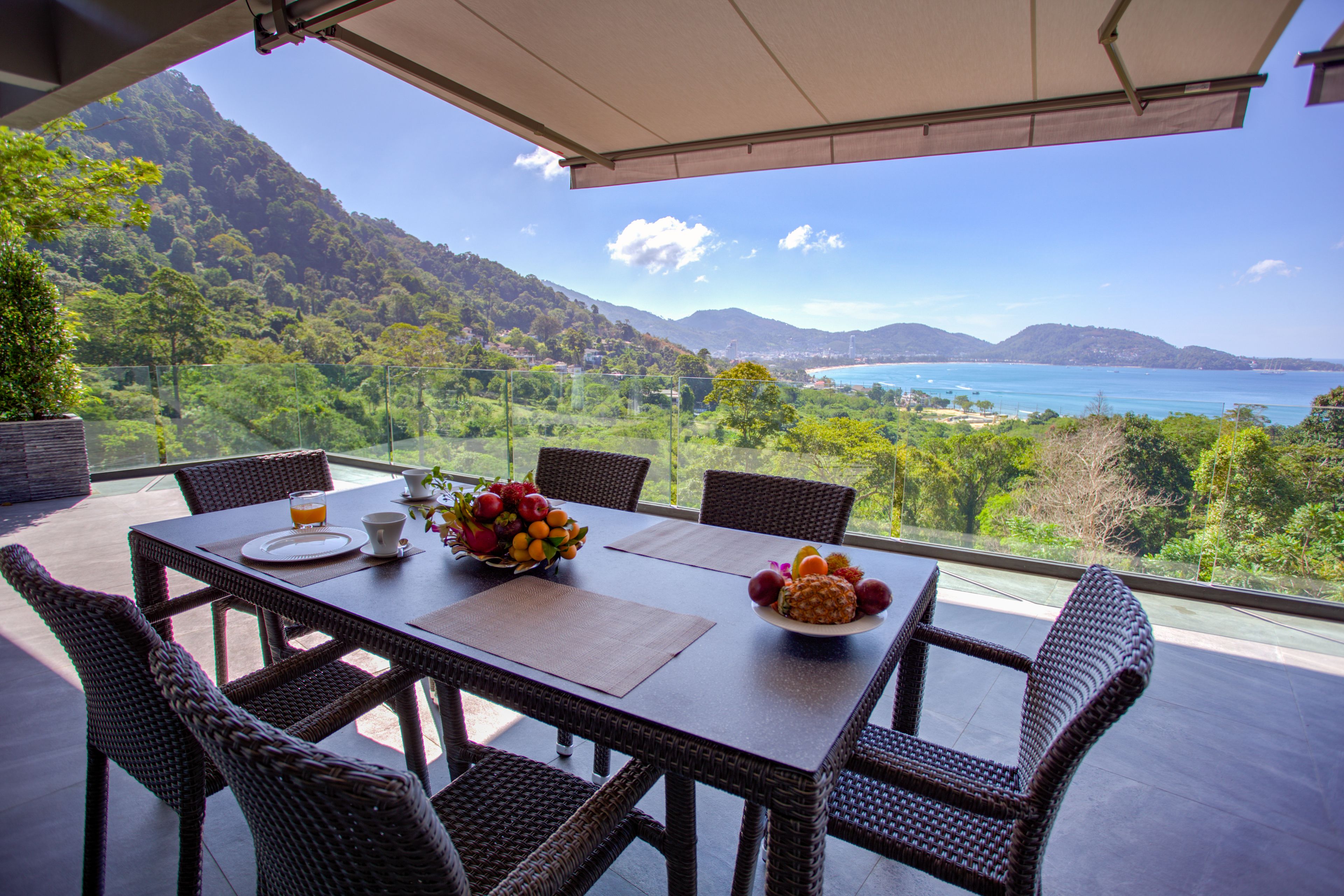 A dining table with wicker chairs, a fruit bowl, and a view of a coastal landscape.