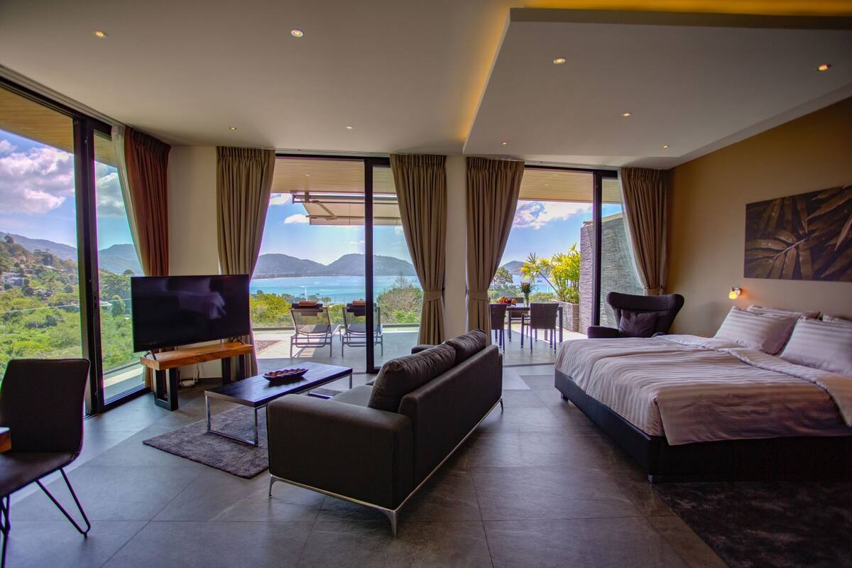 A modern hotel room with a large bed, a sofa, a TV, and a balcony with a view of the sea.