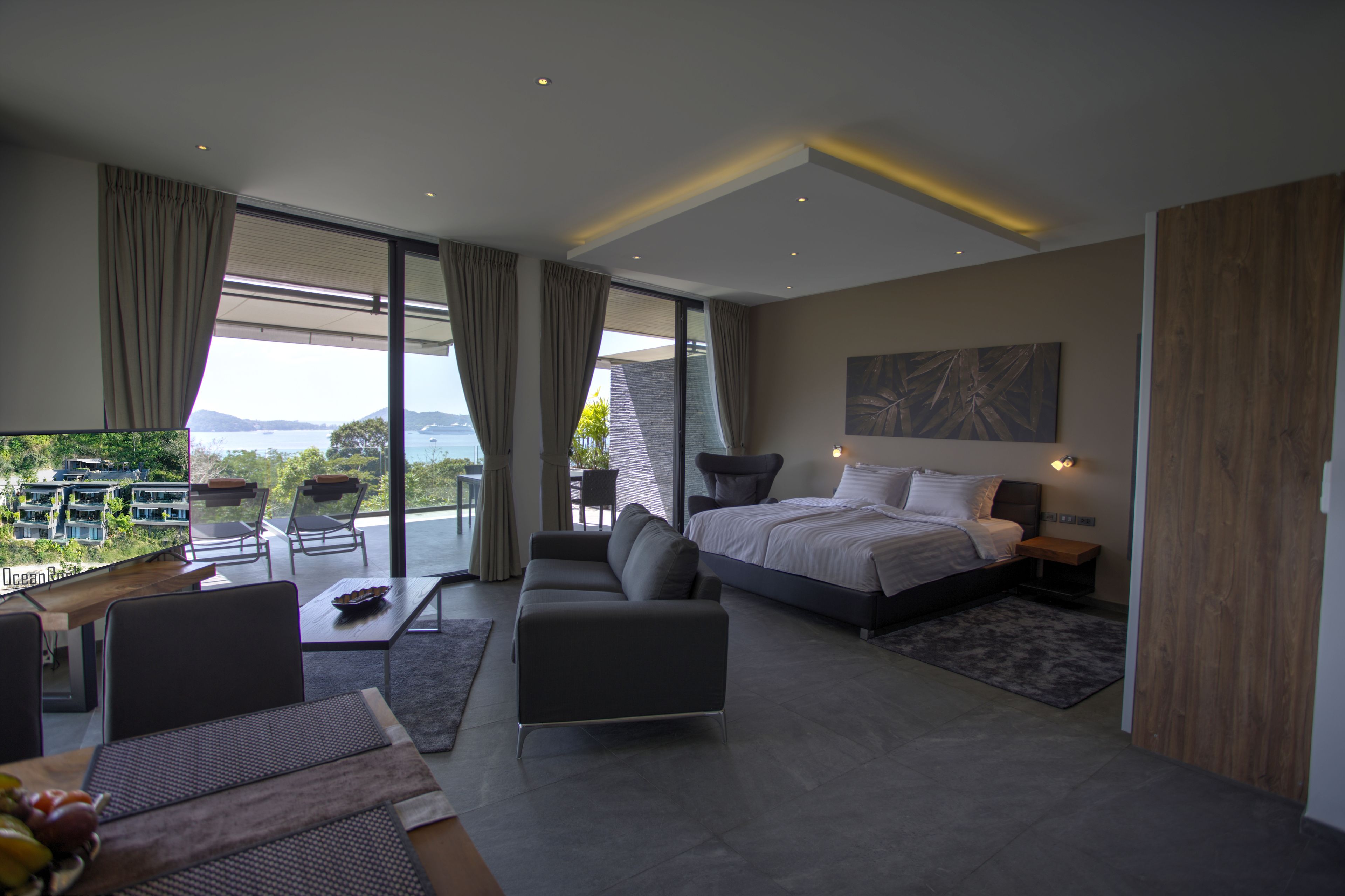 Een moderne slaapkamer met een groot bed, een bank en een eettafel. Er is een balkon met uitzicht op de oceaan en de heuvels.