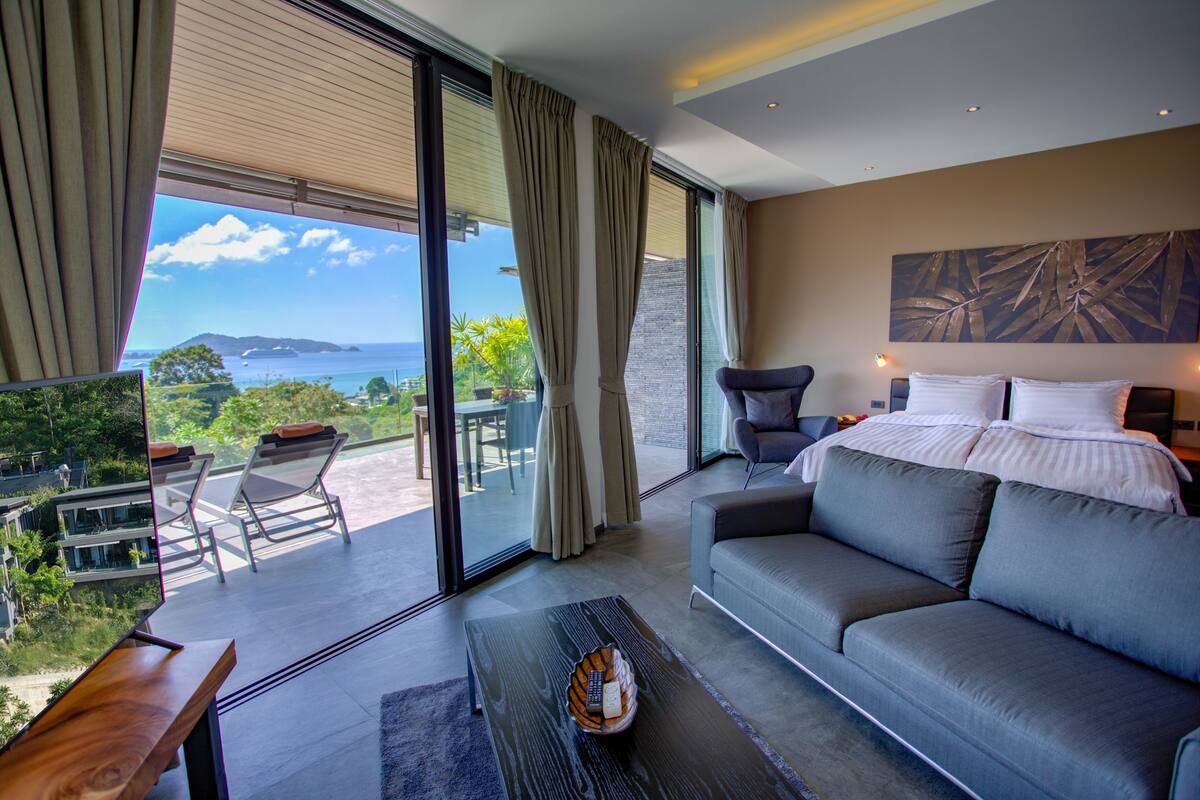 A modern hotel room with a large bed, a sofa, a dining table, and a view of the ocean.