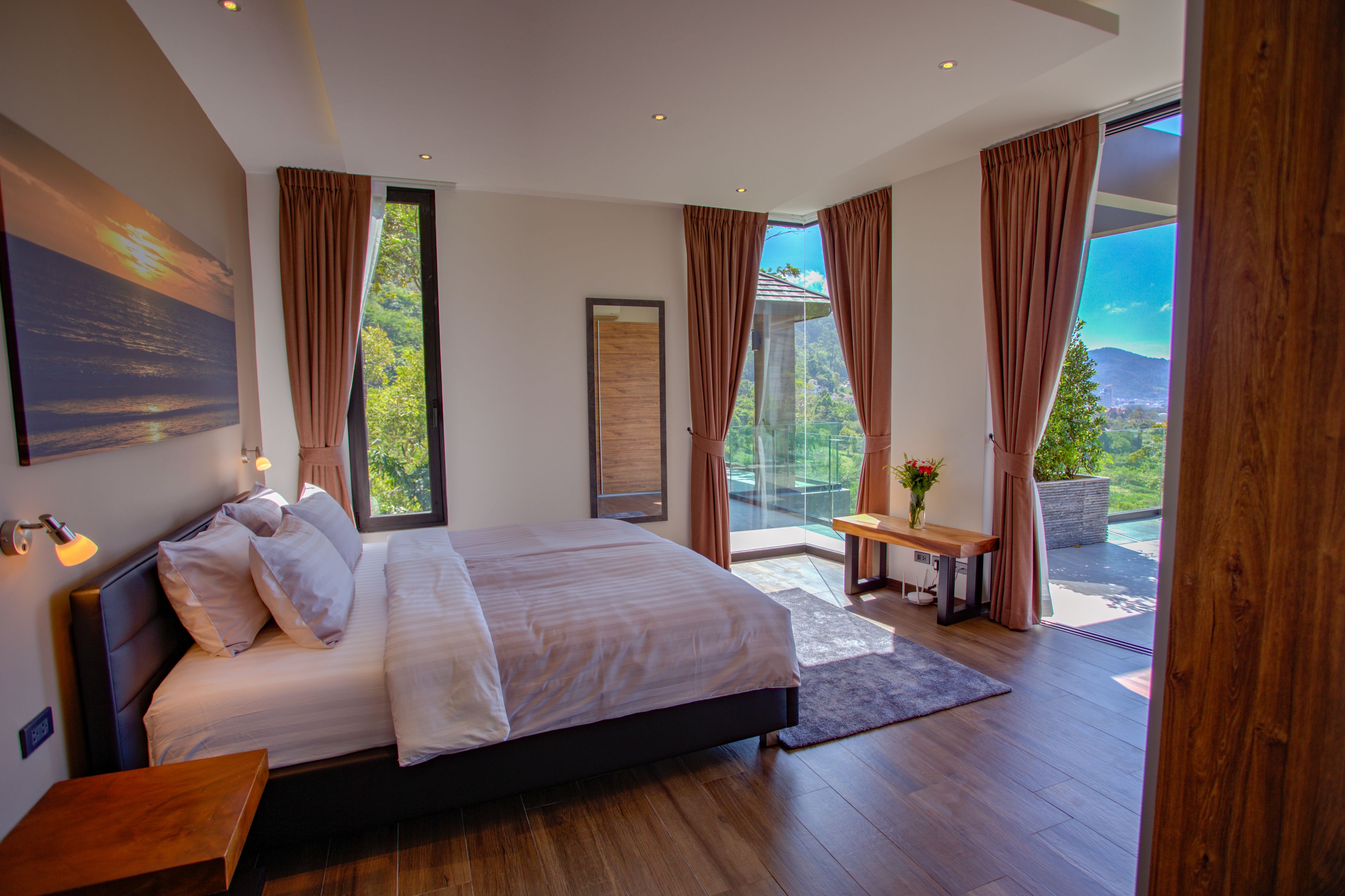 Een moderne slaapkamer met een groot bed, een klein tafeltje met een vaas, en uitzicht op groen en bergen door grote ramen.