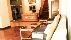 En hotellobby med en receptionsdisk märkt med texten ”HAVENS HOTEL”, en trappa och en sittgrupp med en soffa och fåtöljer.