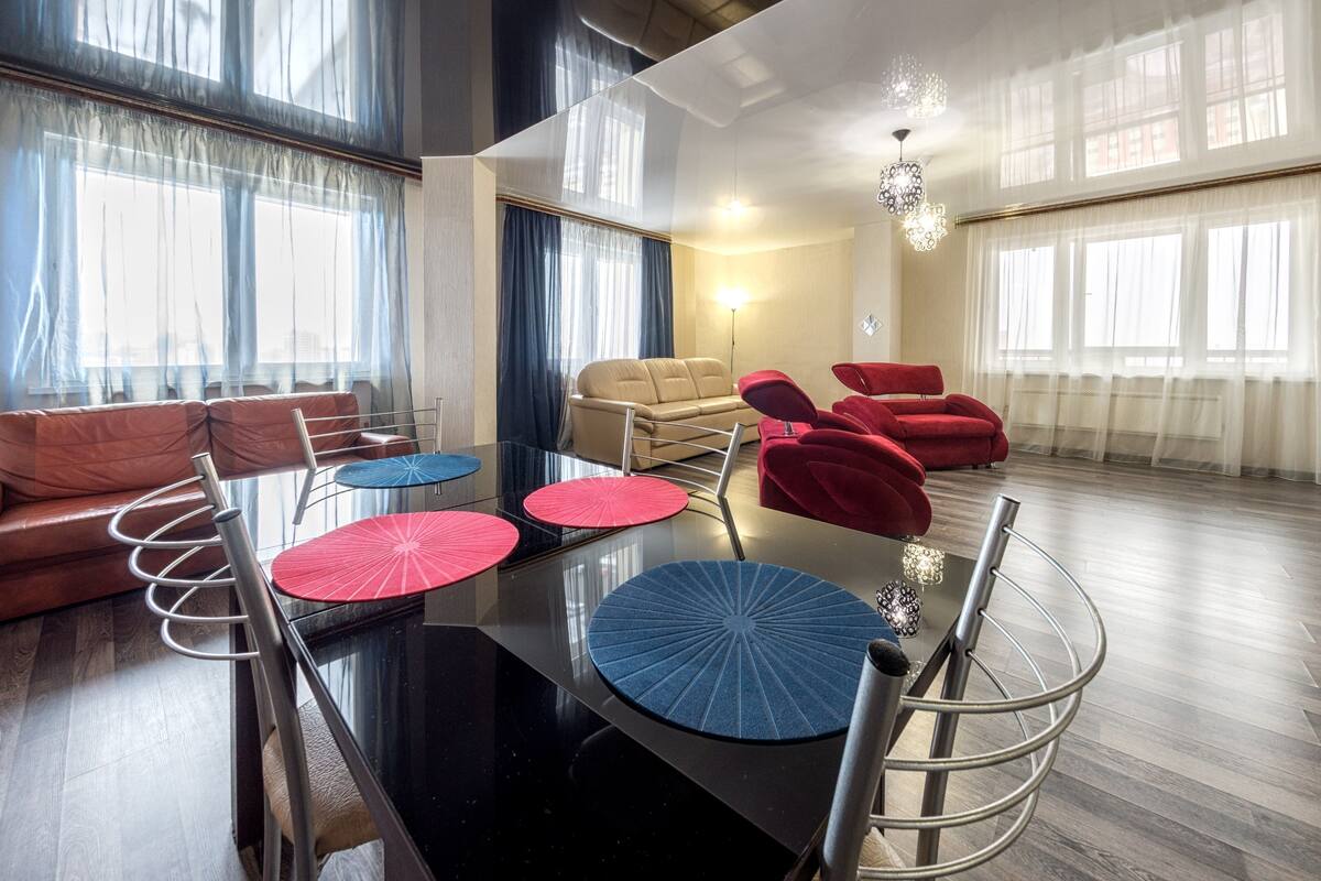 A modern living room with a glass partition, leather sofas, a dining table with red and blue placemats, and a chandelier.