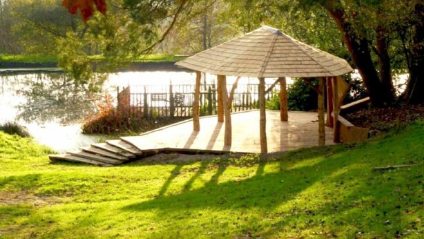A gazebo with a sloped roof, surrounded by grass and trees, near a body of water. - Deers Leap Retreat (South Molton)