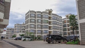A multi-story residential building with balconies, a paved driveway, and parked cars. - Apple Residdence (Syokimau)