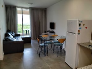 Un espacio habitable compacto con sofá, mesa de vidrio y sillas, pequeña cocina con refrigerador, y balcón con cortinas.