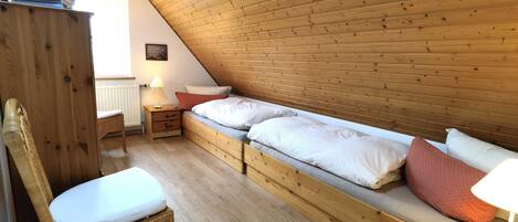 A cozy attic bedroom with a wooden sloped ceiling, a single bed with pillows, a wicker chair, a small wooden table, and a nightstand with a lamp.