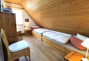 Acogedor dormitorio en ático con techo inclinado de madera, una cama individual con almohadas, una silla de mimbre, una pequeña mesa de madera y una mesita de noche con lámpara.