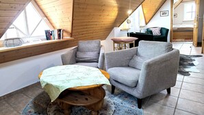 Un acogedor dormitorio tipo loft con techo inclinado, un banco de madera, dos sillones, una pequeña mesa y vista de la habitación contigua.