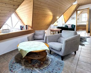 Un acogedor dormitorio tipo loft con techo inclinado, un banco de madera, dos sillones, una pequeña mesa y vista de la habitación contigua.