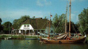 Un edificio tradicional con un letrero que dice 'Raucherhaus', junto a un cuerpo de agua tranquilo con un velero amarrado.