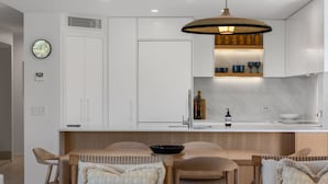 A modern kitchen with white cabinets, a wooden dining table, and a pendant light. - Generous and Comfortable Beachside Abode - See the Ocean - All on one level, lift, pool (SUNSHINE BEACH)