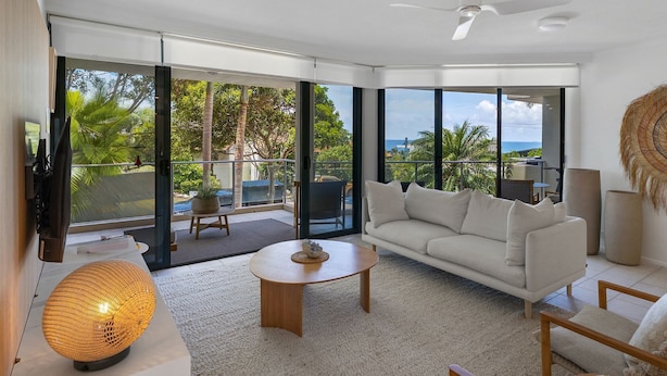 A modern living room with a white sofa, a round coffee table, and a large window offering a view of the ocean and palm trees. - Generous and Comfortable Beachside Abode - See the Ocean - All on one level, lift, pool (SUNSHINE BEACH)