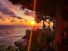 A sunset view over the ocean with a wooden deck and tropical vegetation.