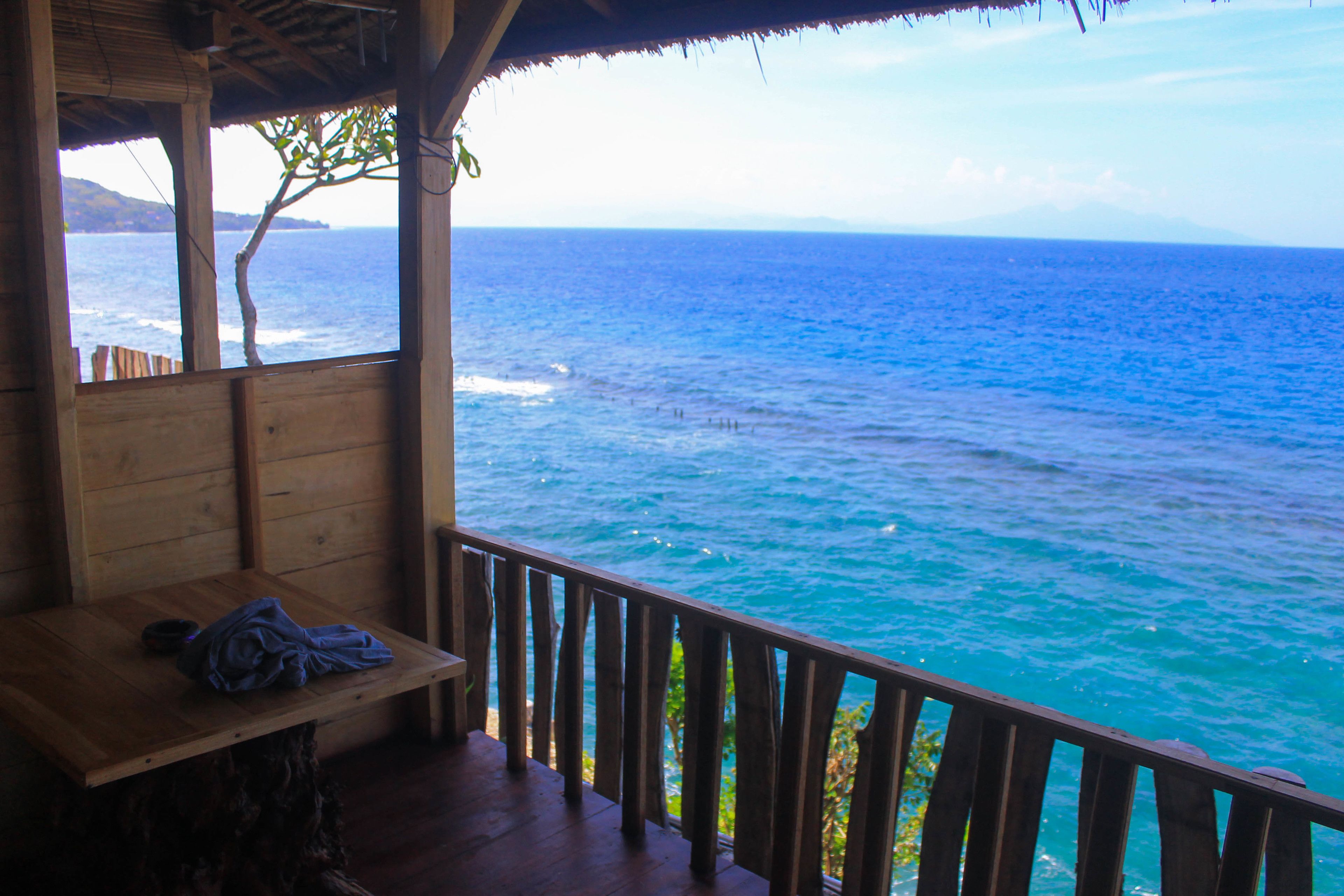 A wooden deck with a bench and a table, overlooking a turquoise sea with waves.