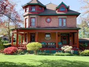 A two-story house with a wrap-around porch, a front door with a wreath, and a small front yard with a lawn and shrubs.