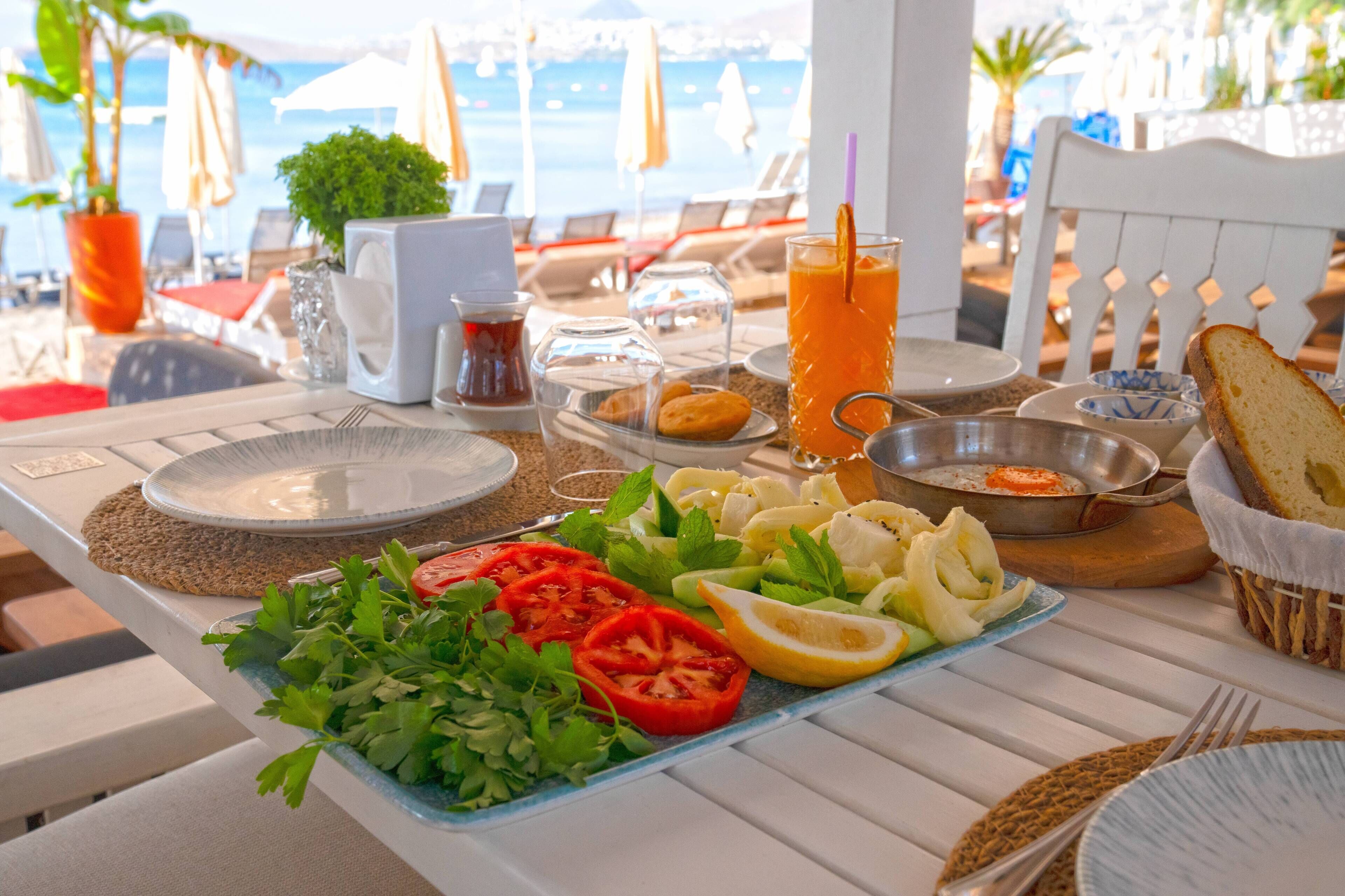 Une table dressée avec une variété d’aliments, notamment des légumes frais, du pain et une boisson, avec une vue sur la mer en arrière-plan.