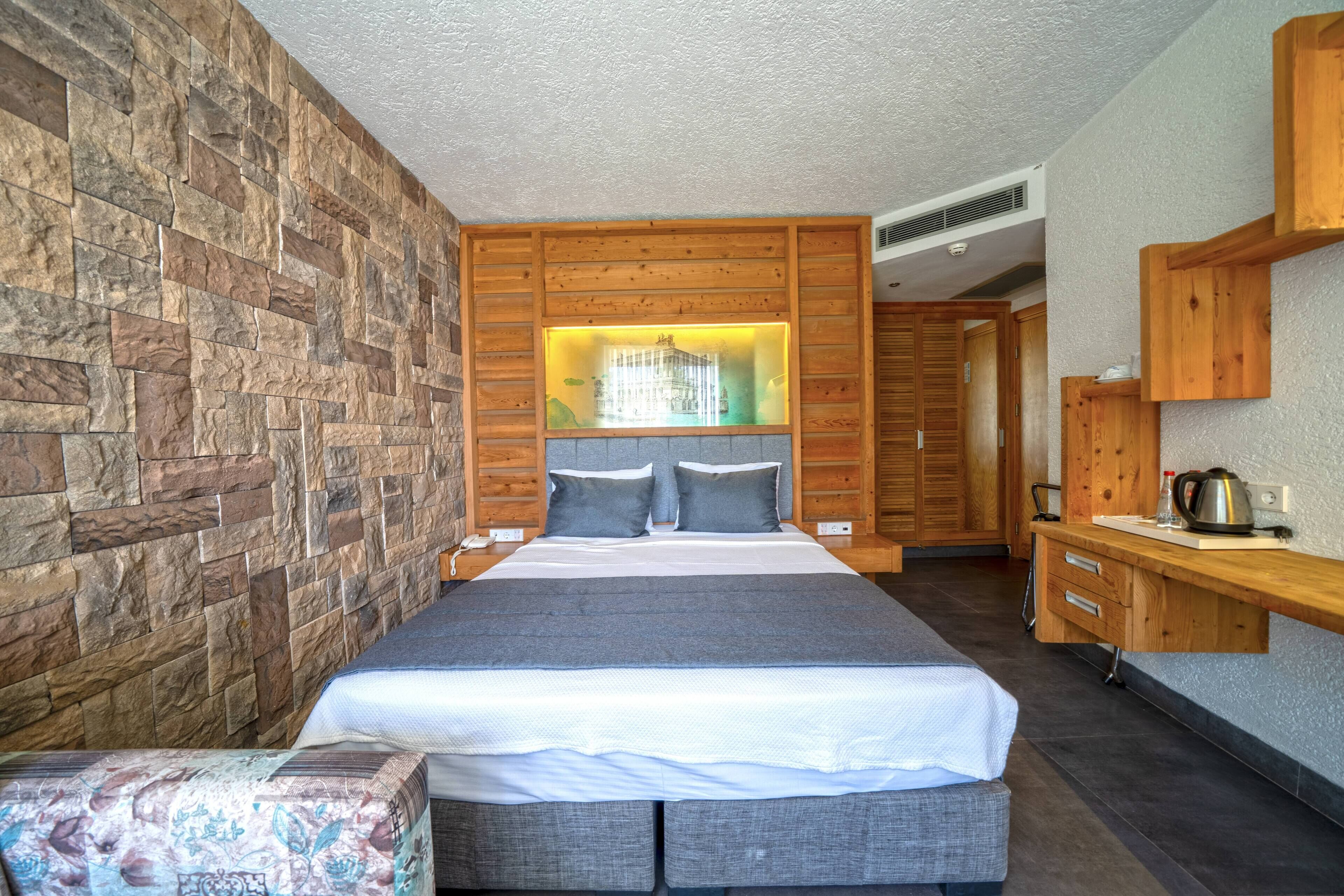 Un dormitorio moderno con pared de piedra, una cama grande con ropa de cama gris, un cabecero de madera y una mesita de noche con hervidor.