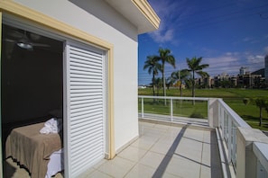 A balcony with a view of palm trees and a grassy area, a bed with a white cover, and a closed sliding door.