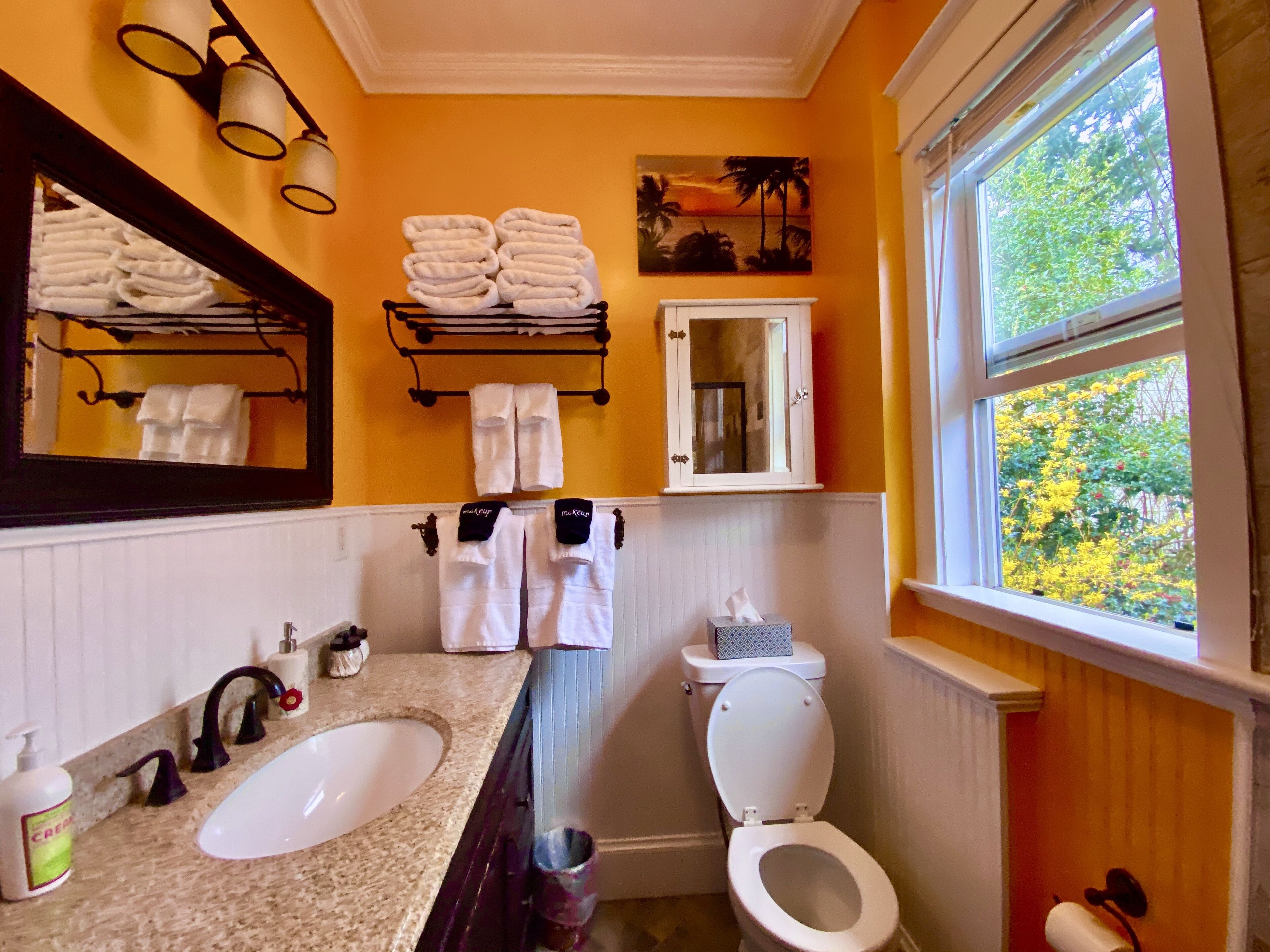 A bathroom with a sink, toilet, and a window with a view of trees. There are towels on a rack and a painting on the wall.