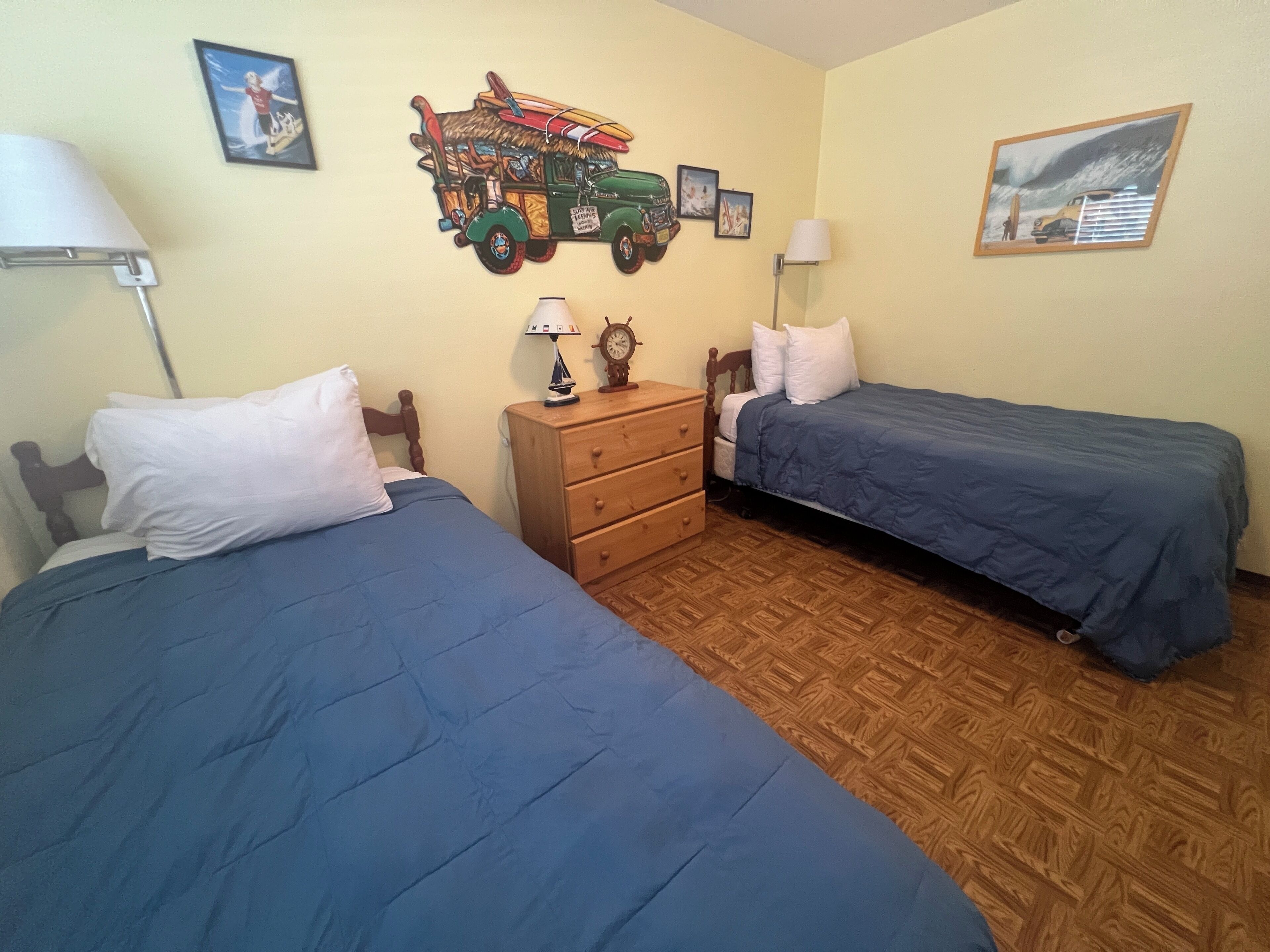 Une chambre avec deux lits, une commode en bois et un mur décoré de planches de surf et d’une image de camion.