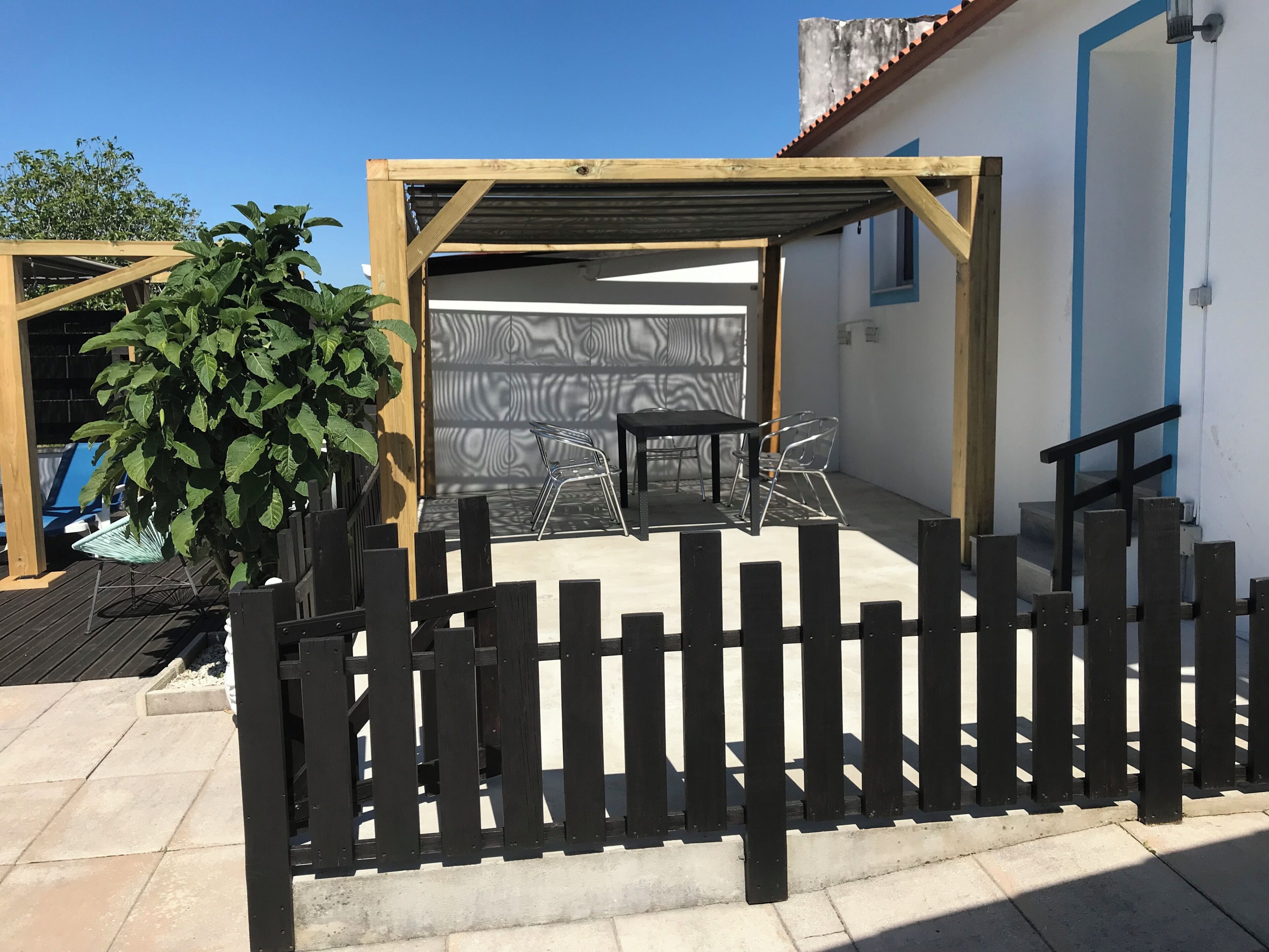 Ein überdachter Außenbereich mit einer Holz-Pergola, einem schwarzen Metallzaun und einem Blumentopf.