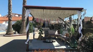 Un espace extérieur aménagé, abrité et doté d’un canapé et de coussins, entouré de palmiers et d’une clôture décorative.