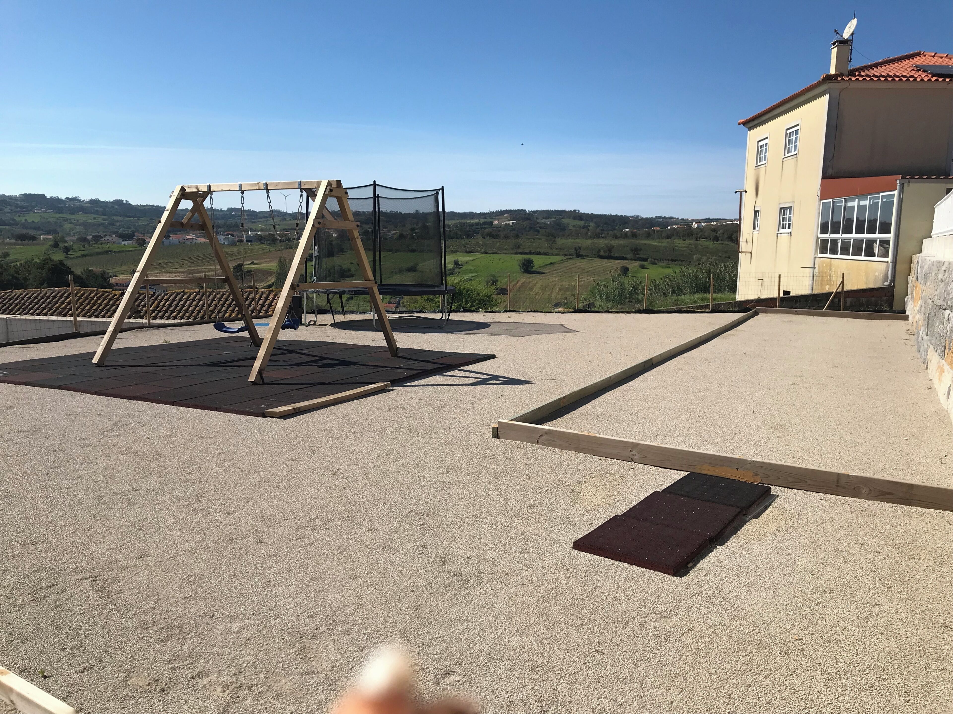 A rooftop with a swing set, a trampoline, and a building with a red roof in the background.