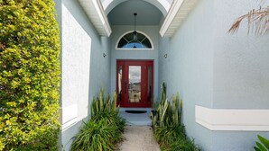 A narrow pathway leading to a red door with a white porch, flanked by green plants and a small table.