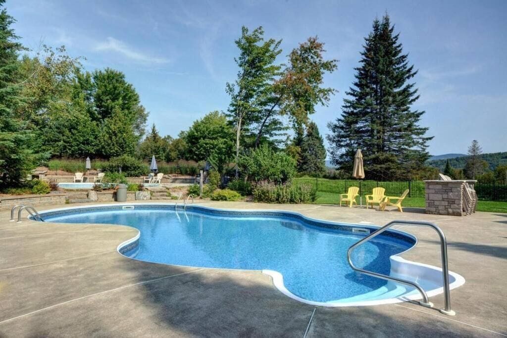 A swimming pool with a curved edge, surrounded by a concrete deck. There are chairs and a small stone structure in the background.