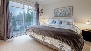 Une chambre avec une grande fenêtre, un lit recouvert d’une couvre-lit à motifs, une table de chevet avec une lampe et une œuvre d’art accrochée au mur.