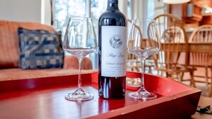 Une bouteille de vin sur un plateau, accompagnée de deux verres, est posée sur une table en bois dans une pièce où se trouvent un canapé rouge et, au fond, un coin repas.