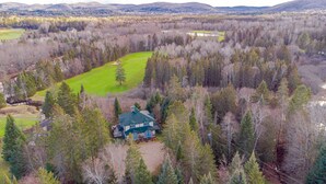 Une grande maison entourée d’une forêt dense, avec une pelouse bien entretenue et un petit étang.