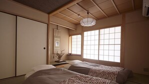 A traditional Japanese room with tatami flooring, two beds with patterned bedding, a shoji screen window, and a hanging light fixture.