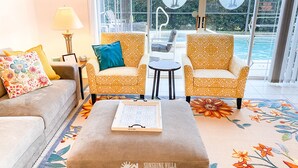 A sunlit living room with a sofa, two patterned armchairs, a coffee table, and a view of a pool and greenery outside.