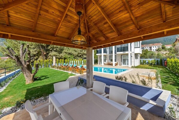 A covered outdoor seating area with a pool and a view of a modern building. - 4 bedroom luxury villa sardunya in ölüdeniz fethiye (fethiye ölüdeniz)