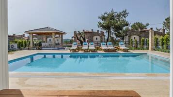 En swimmingpool med liggestole, en pergola og stenhuse i baggrunden.