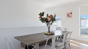 A dining area with a wooden table and white chairs, a vase with flowers, and a view of a house through a window.