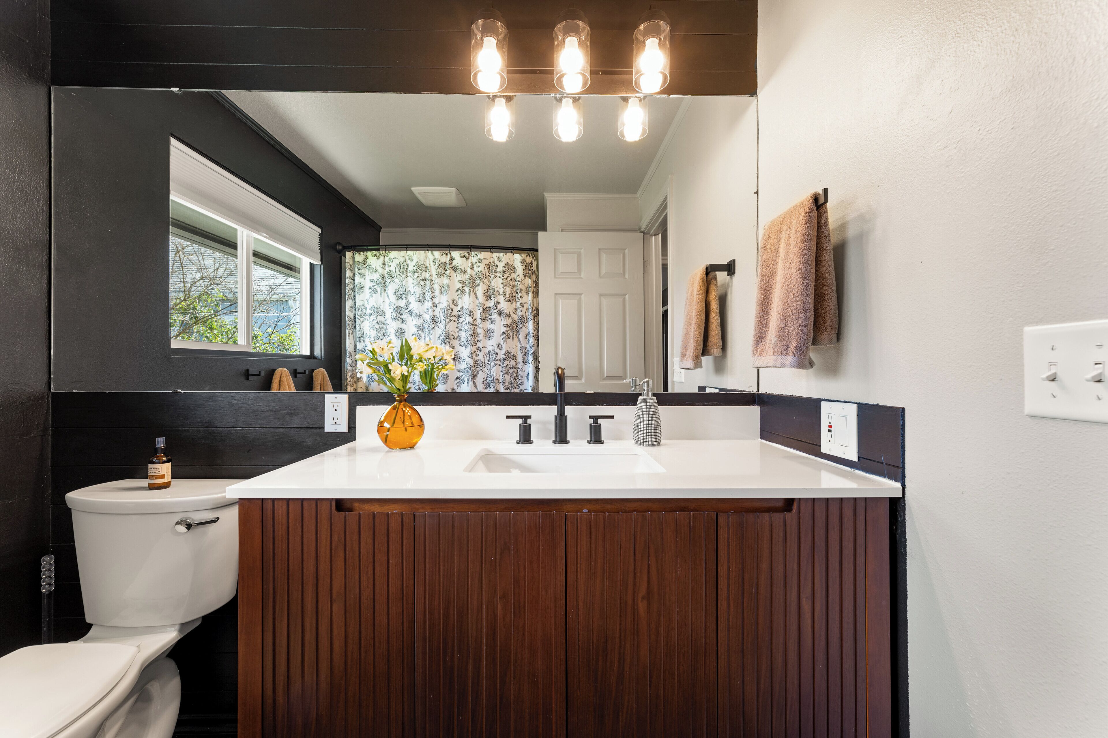 A modern bathroom with a large wooden vanity, a white sink, and a toilet. There is a window with a view of trees and a door with a mirror above it.